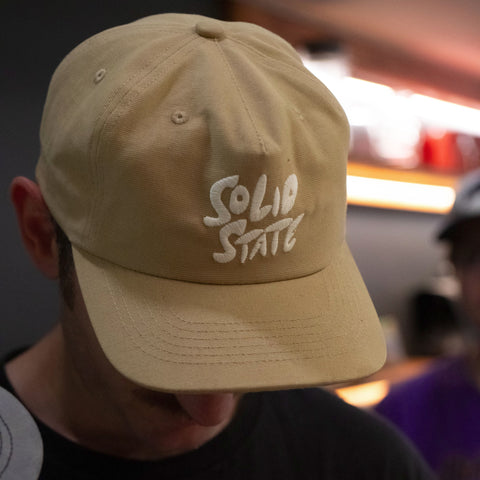A man wearing a beige baseball cap with 'Solid State' embroidered in white on the front, photographed in a dimly lit indoor setting.